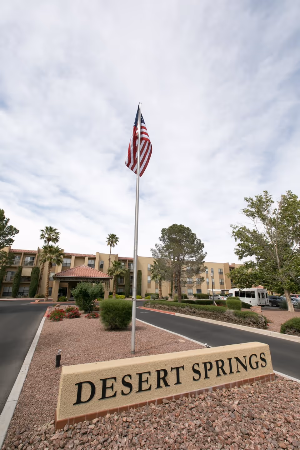 Exterior view of Desert Springs Senior Living facility with a large American flag on a flagpole in the center. The building is a multi-story structure with beige walls and a red-tiled roof. There are palm trees and other greenery around the entrance, and a driveway leading up to the building. A sign in the foreground reads 'DESERT SPRINGS'.