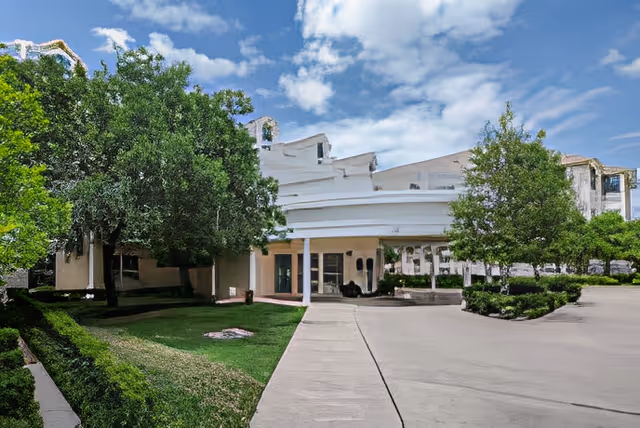 Exterior view of a senior living facility building with a covered entrance, surrounded by green trees and bushes under a partly cloudy blue sky.