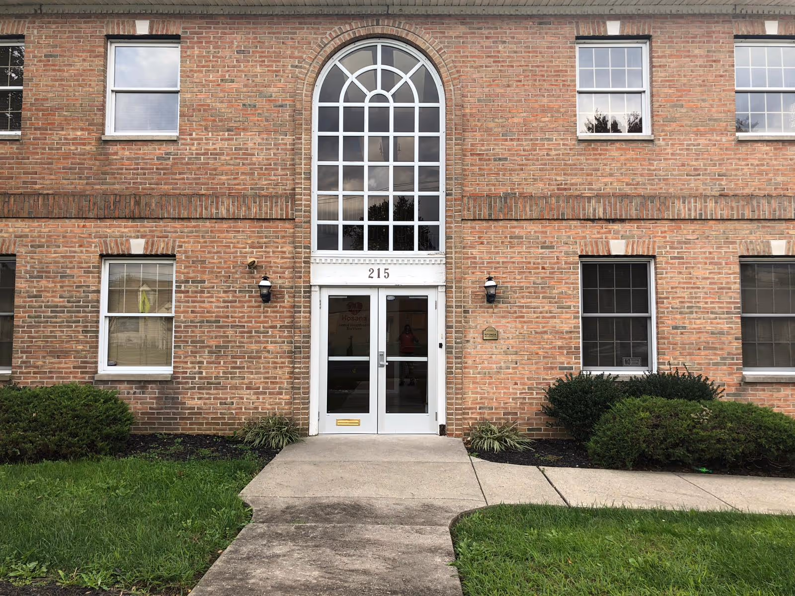 Brick facility entrance with arched window above and manicured shrubs.