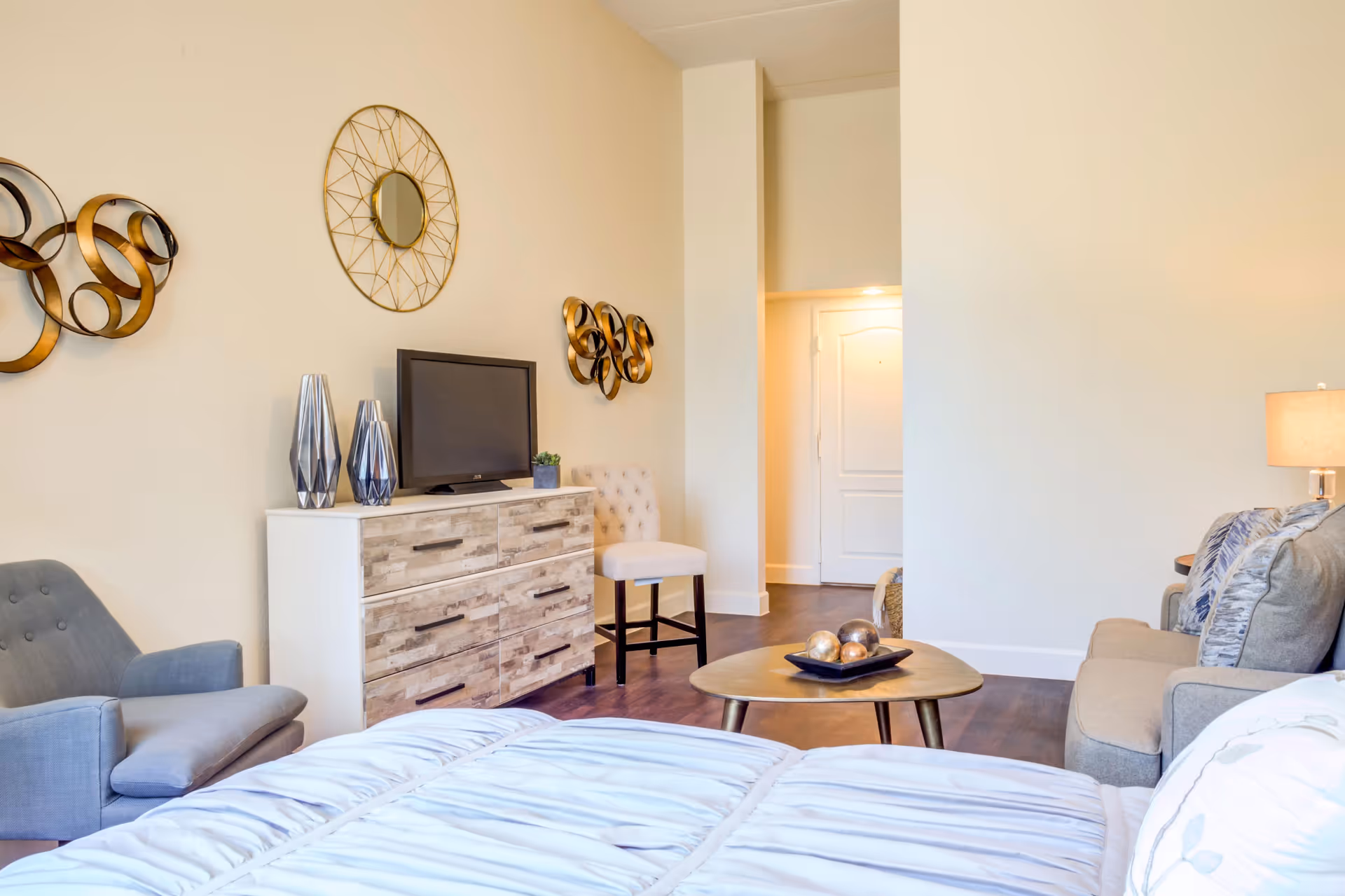 A cozy senior living room with a gray armchair, a wooden dresser with a small TV and decorative vases, a round coffee table with decorative balls, a beige sofa with cushions, and wall art including a round mirror and metal sculptures. The room has light-colored walls and wooden flooring, with a doorway leading to another area.