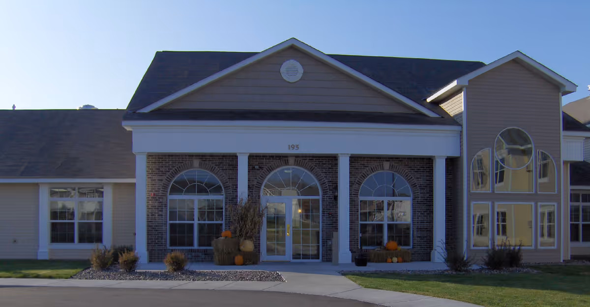 Front exterior view of a senior living facility building named Traditions of Owatonna, featuring a brick and siding facade with large arched windows, a central entrance door, and autumn decorations including pumpkins and hay bales near the entrance.