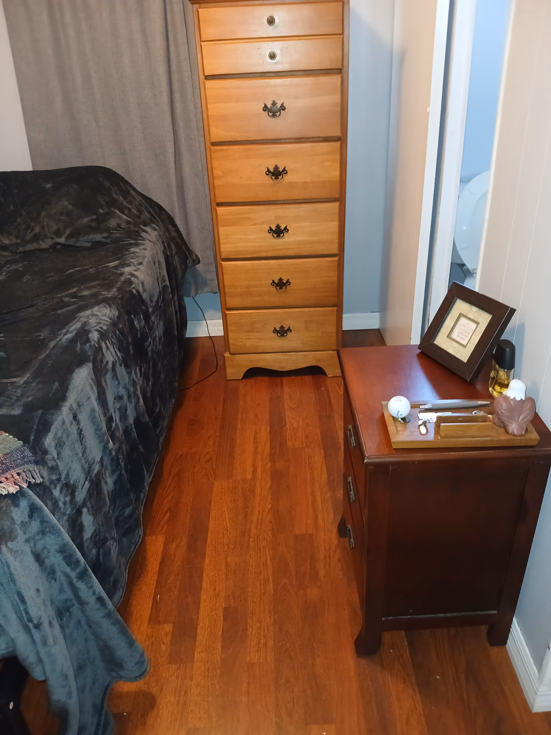 Bedroom scene with bed, tall dresser, nightstand, and framed items on a hardwood floor.