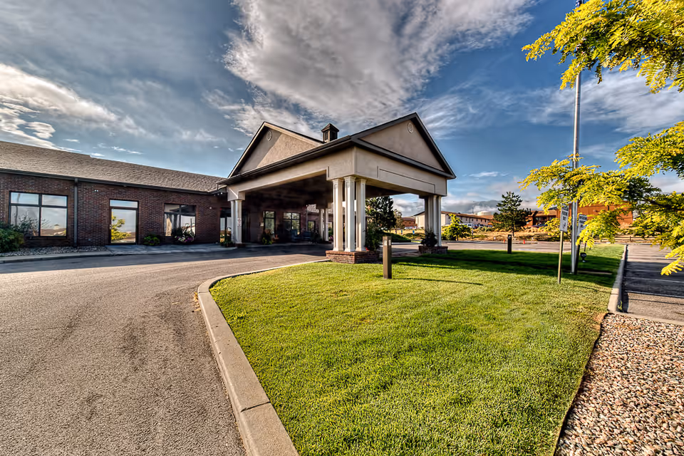 Front entrance of a single-story brick senior living building with a covered porte-cochere, driveway, and well-kept lawn under a partly cloudy sky.