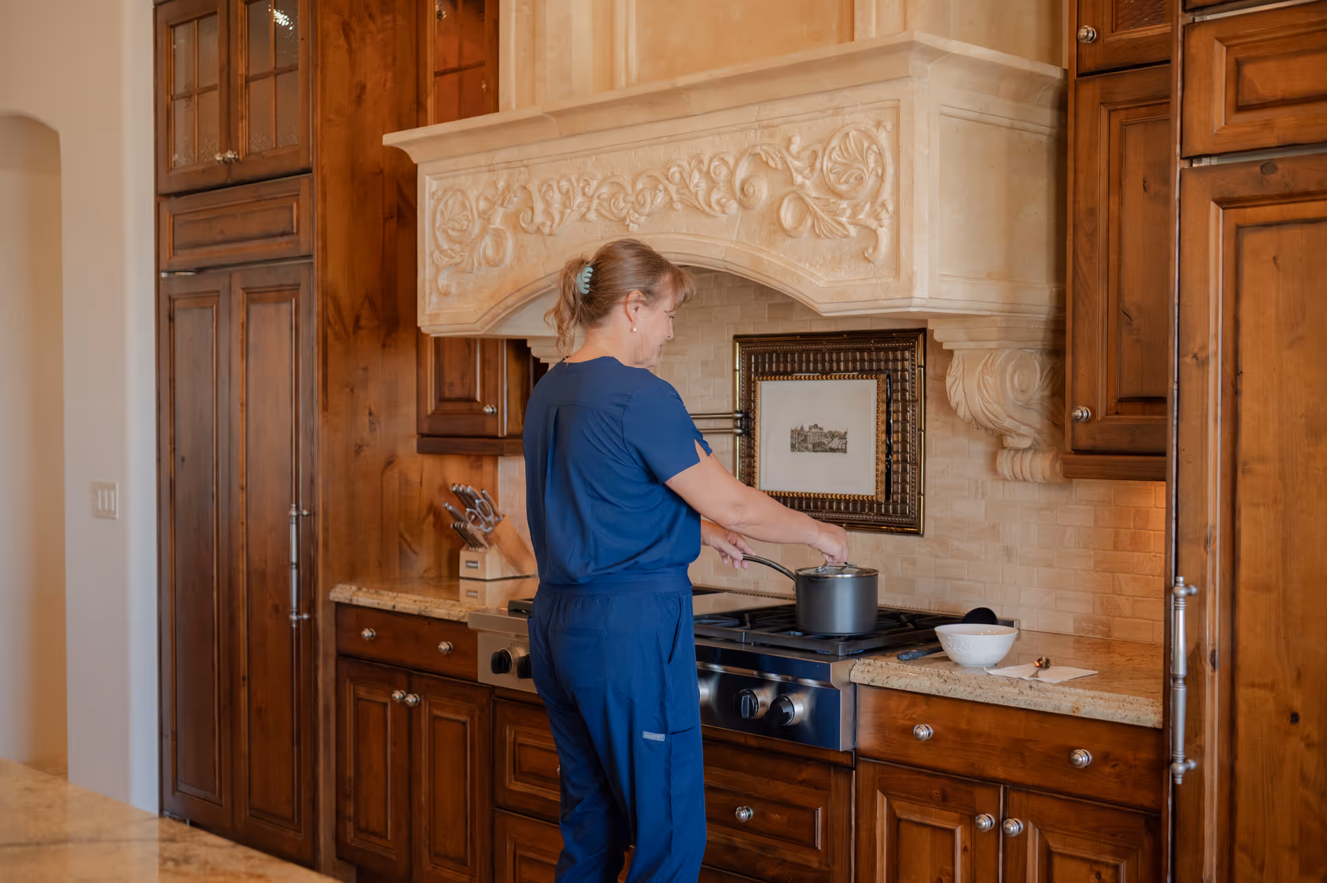 Caregiver in blue scrubs stirs a pot on the stove in a wooden kitchen.