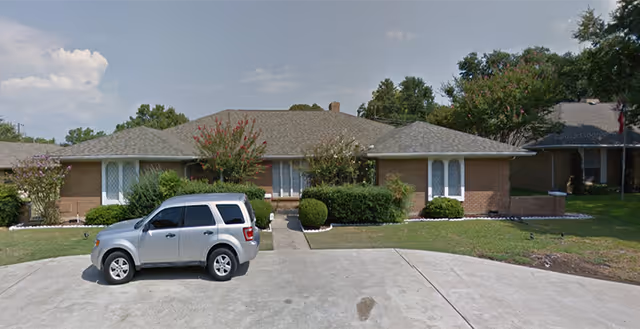 Single-story brick senior living facility front with a silver SUV parked on the driveway and shrubs.
