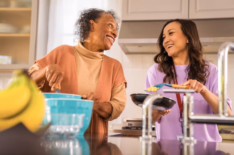 Two women, resident and caregiver, share a smile while cooking together in a bright kitchen.