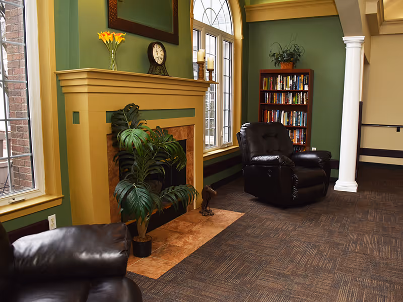A cozy living room area with green walls and yellow trim, featuring a fireplace with a clock and flowers on the mantel. There are two black leather recliners, a tall bookshelf filled with books, a large window with an arched top, a potted plant in front of the fireplace, and a white decorative column near the bookshelf.