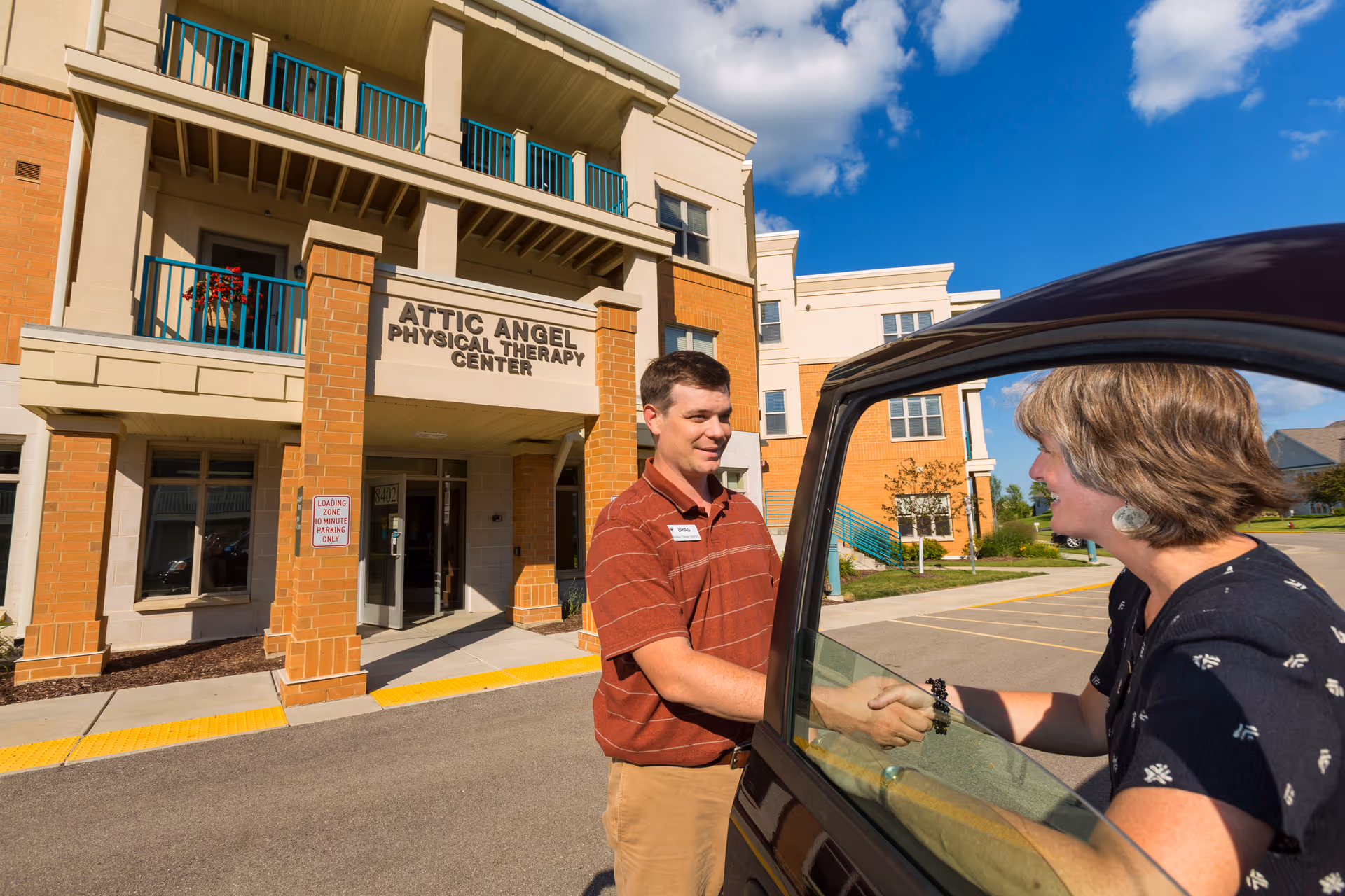 Two people shake hands through a car window in front of Attic Angel facility entrance.
