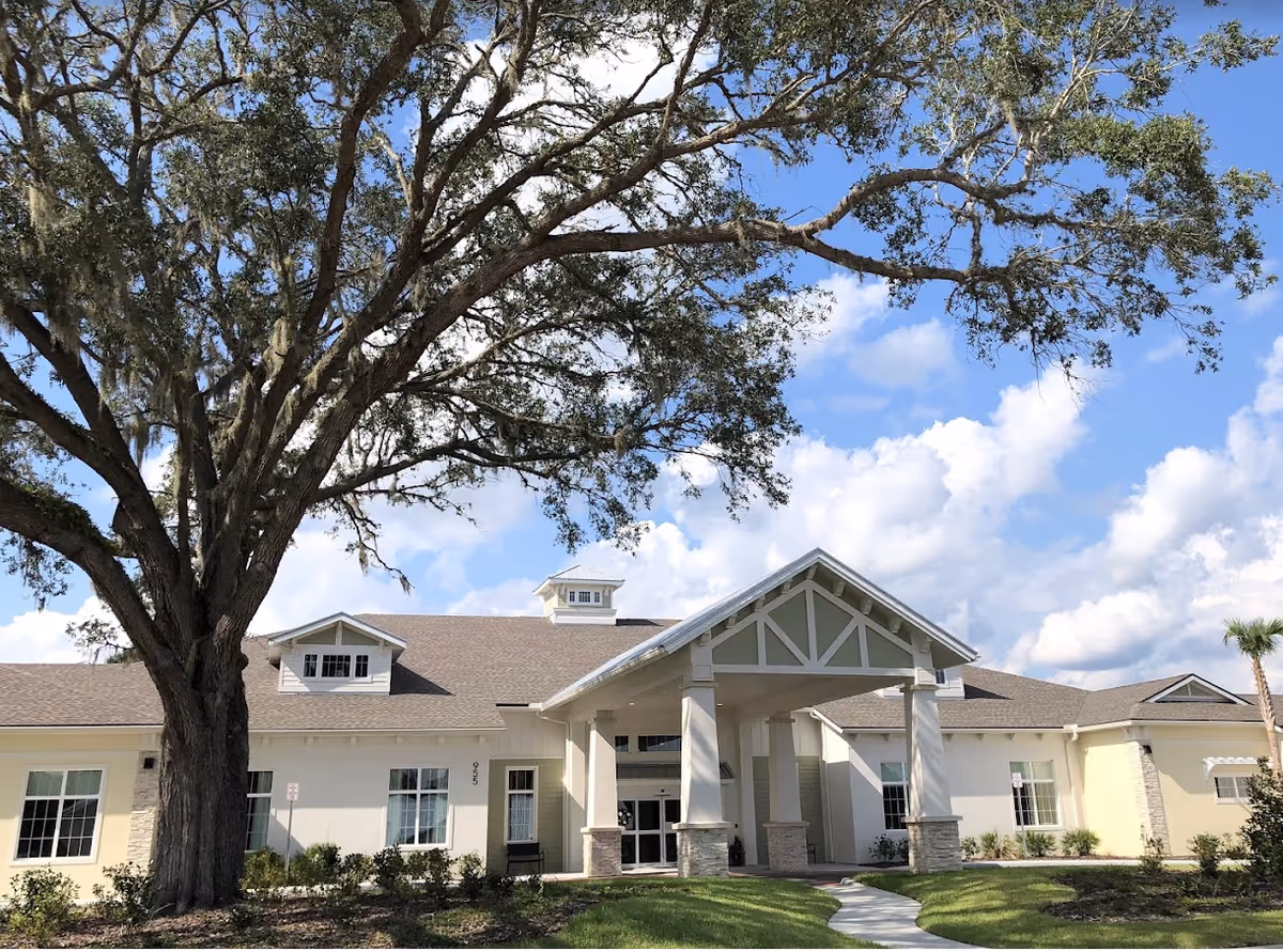 Front exterior of a single-story beige senior living building with a covered entrance and a large oak tree in the foreground under a blue sky.