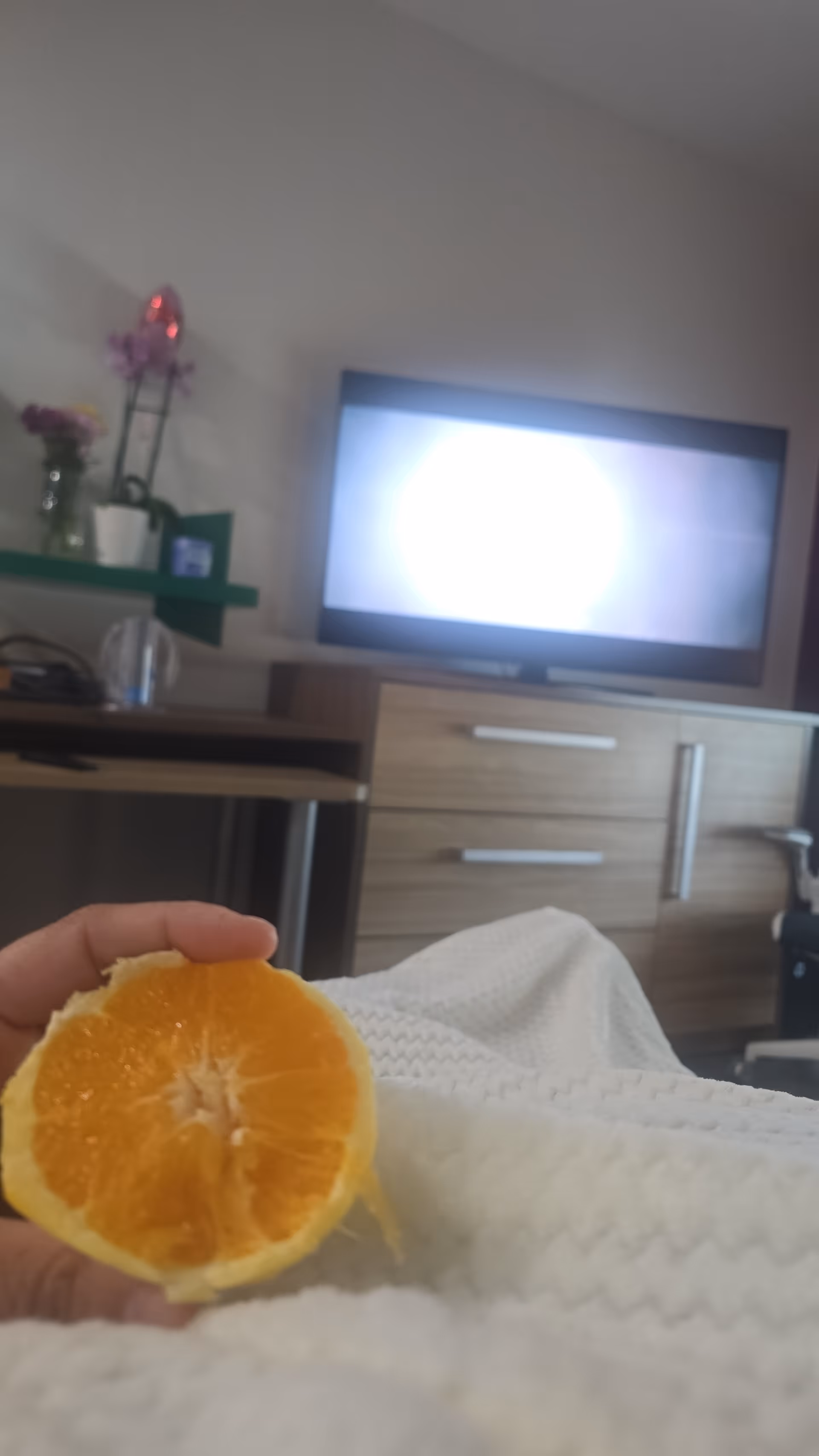 A hand holds half an orange on a white blanket in a bedroom with a TV.