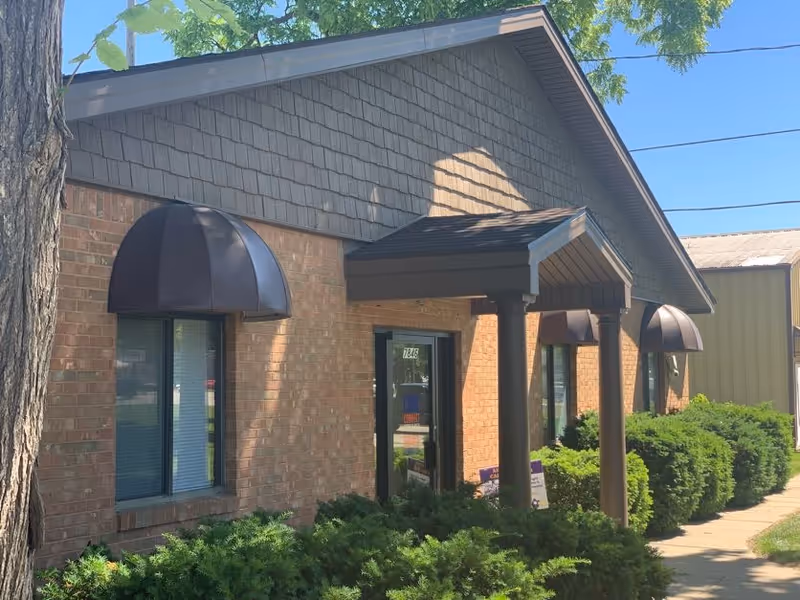 Brick senior care building with arched awnings, glass doors, and green shrubs along a walkway.