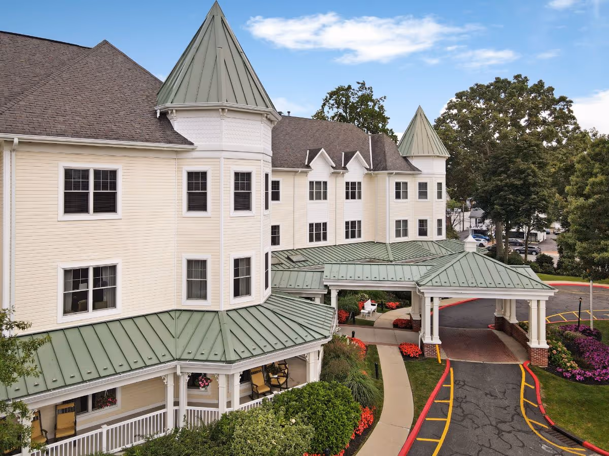Front exterior of a multi-story cream-colored senior living building with green metal roofs, turrets, a covered porte-cochere, driveway, and landscaped entrance.