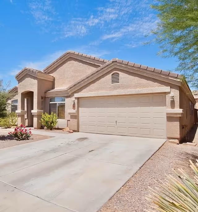 Single-story beige stucco house with a two-car garage, driveway, and desert landscaping under a blue sky.