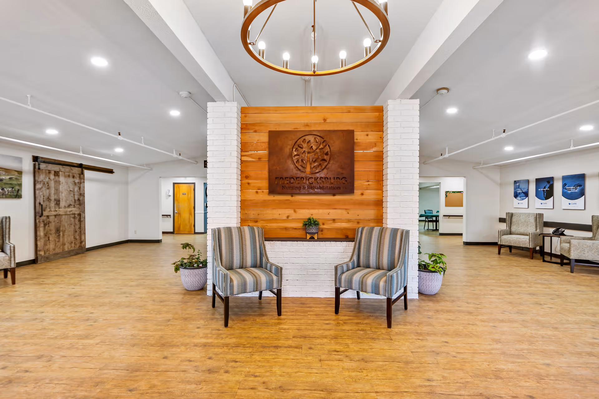 A spacious and well-lit common area in Fredericksburg Nursing and Rehabilitation featuring two striped armchairs in front of a wooden and white brick wall with a sign displaying the facility's name and logo. The room has wooden flooring, potted plants, a rustic sliding barn door on the left, additional seating on the right, and framed pictures on the far wall.