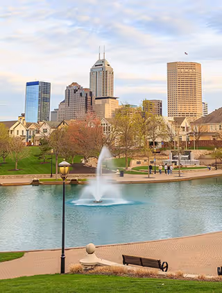 Outdoor park area with a pond, fountain, benches, and a distant city skyline.