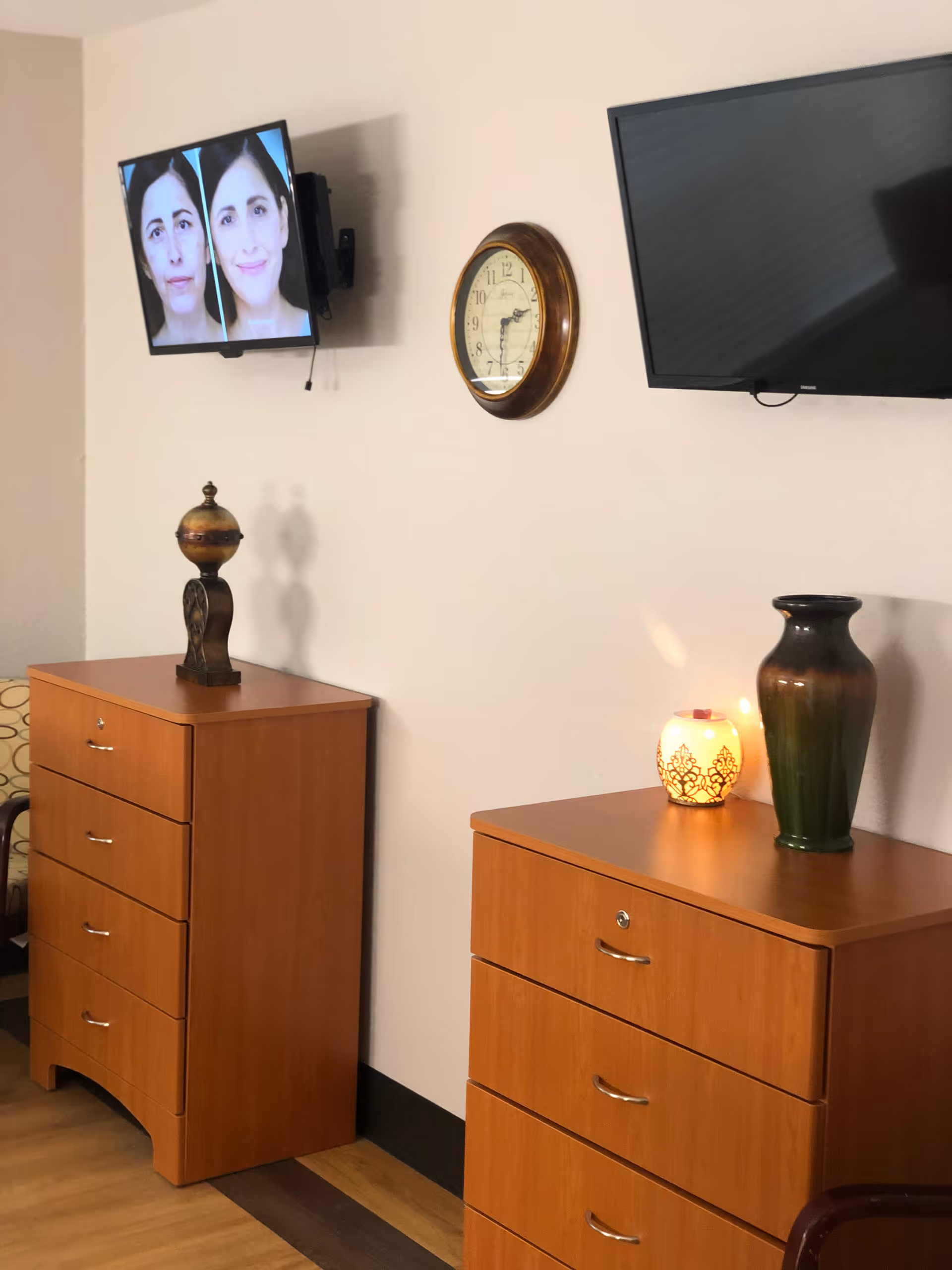 Interior view of a room with two wooden dressers, each topped with decorative items including a clock, a vase, and a lit candle holder. Two wall-mounted televisions are visible above the dressers, one displaying a split image of a woman's face before and after a treatment.