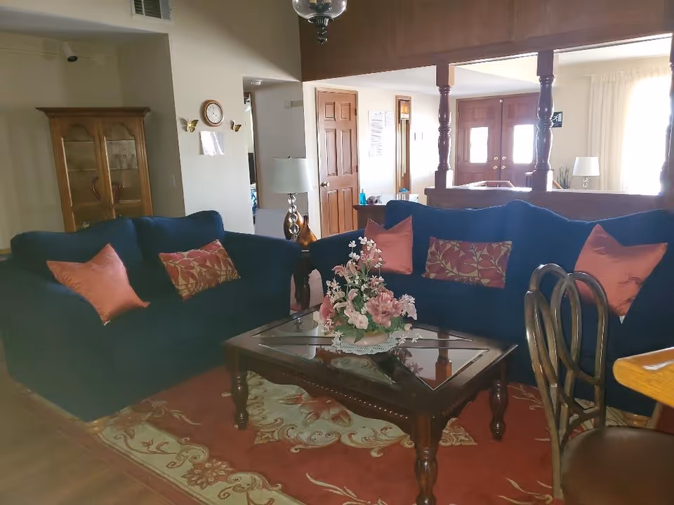 A cozy living room with two dark blue sofas adorned with red and patterned cushions, a wooden coffee table with a glass top and a floral centerpiece, a wooden cabinet with glass doors, a floor lamp, and a wooden dining chair partially visible. The room has wooden doors and a bright window with white curtains in the background.