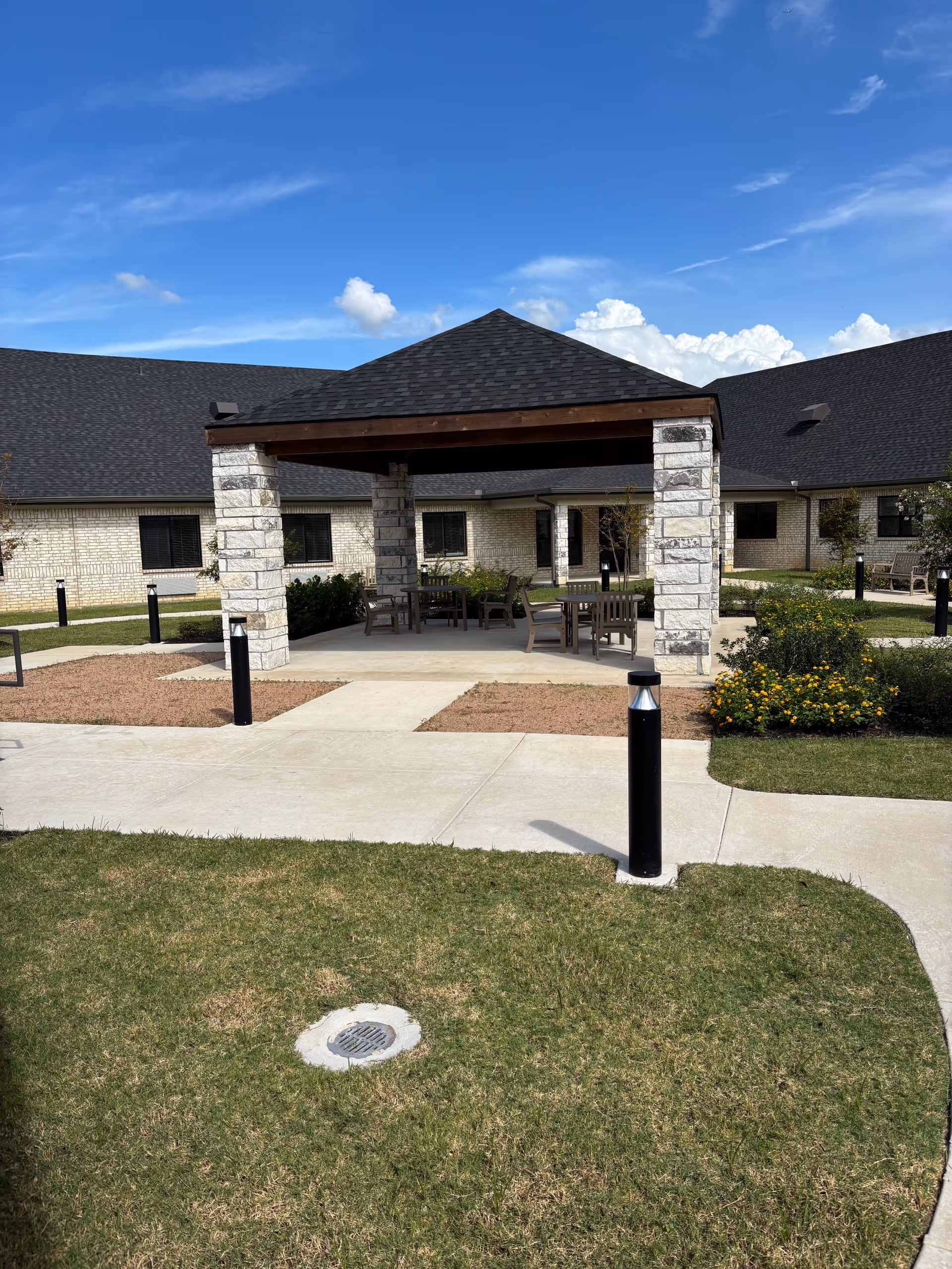 Outdoor seating pavilion with stone columns, tables, and surrounding walkway in a senior living campus.
