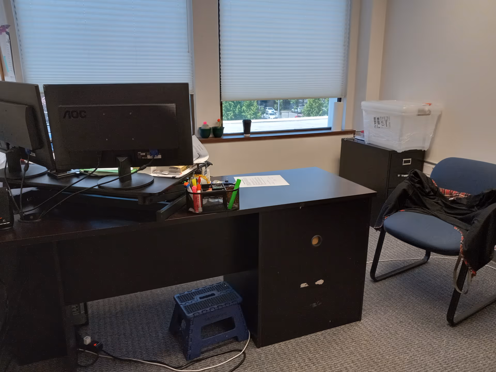 Dark office desk with dual monitors, supplies, and a chair near a windowed wall.