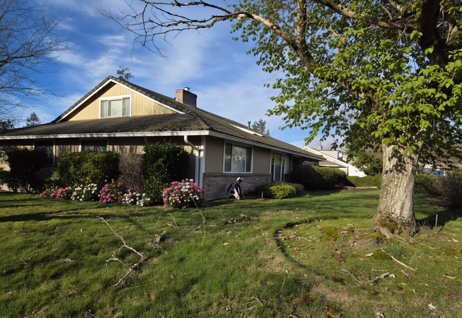 Front exterior view of a single-story home with lawn, trees, and flower beds.