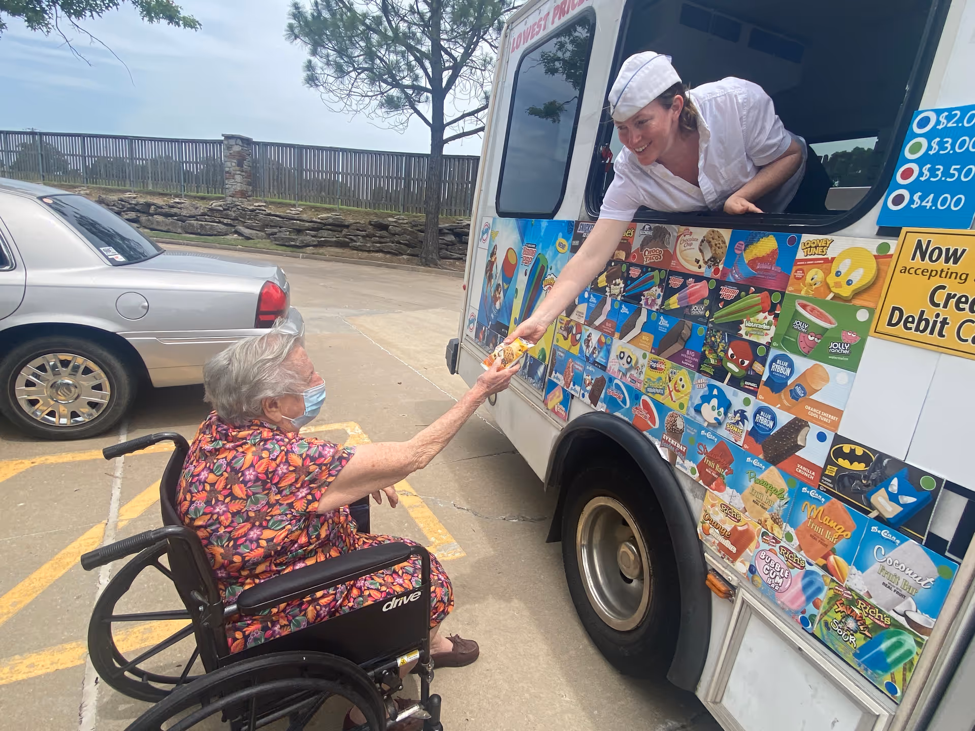 An elderly woman in a wheelchair wearing a floral dress and a face mask is receiving an ice cream from a vendor inside an ice cream truck. The vendor is smiling and handing the ice cream through the truck window. The truck is decorated with colorful images of various ice cream treats and prices. A silver car is parked nearby, and there is a stone wall and tree in the background.