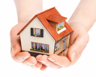 Hands cradle a small model house with a red roof and blue shutters.