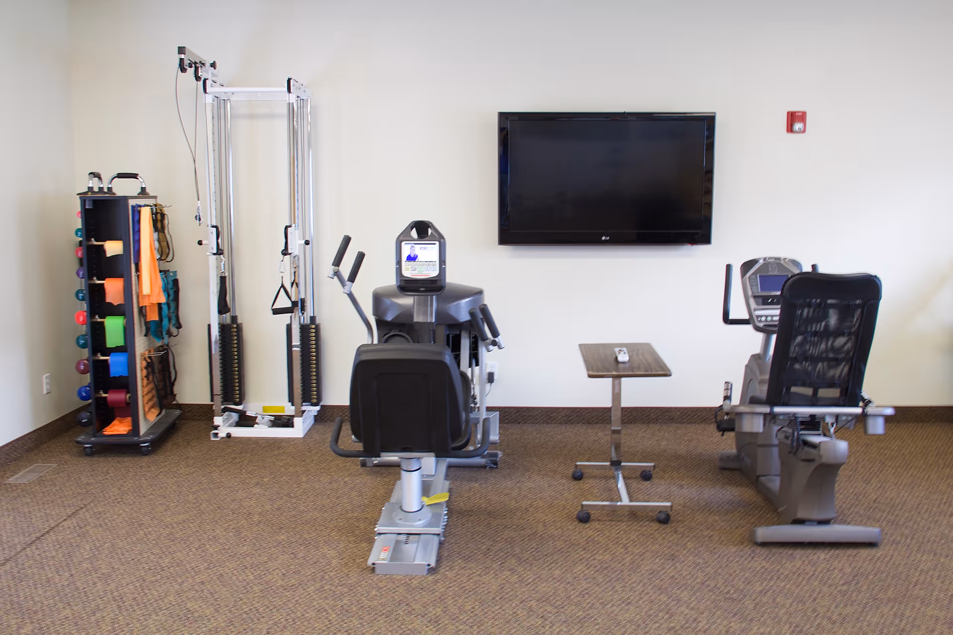 Fitness room with exercise machines and a wall-mounted TV in a care facility.