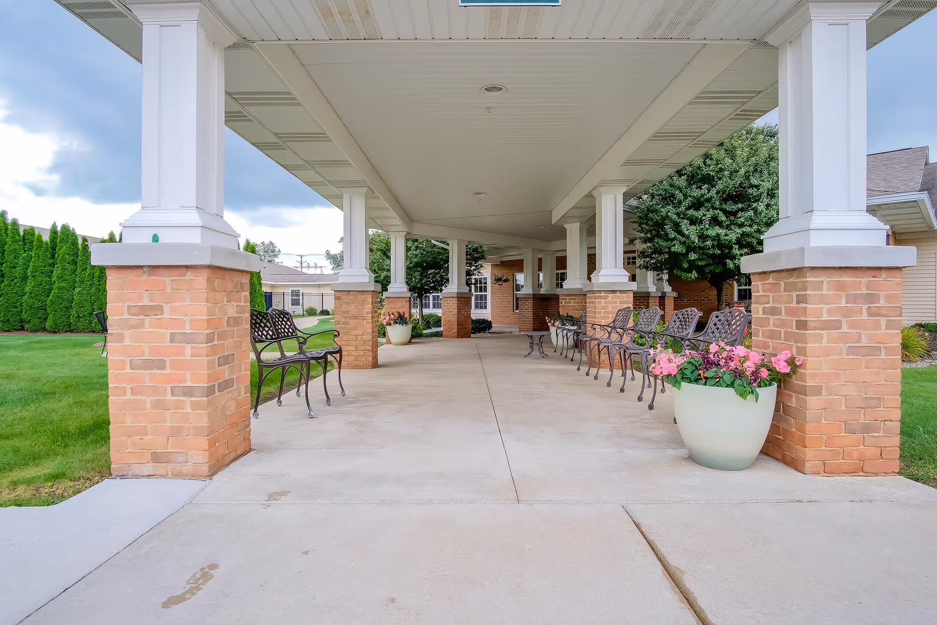 Covered outdoor walkway with brick pillars and white columns, lined with metal benches and large flower pots containing pink flowers. Green grass and trees are visible on either side under a cloudy sky.