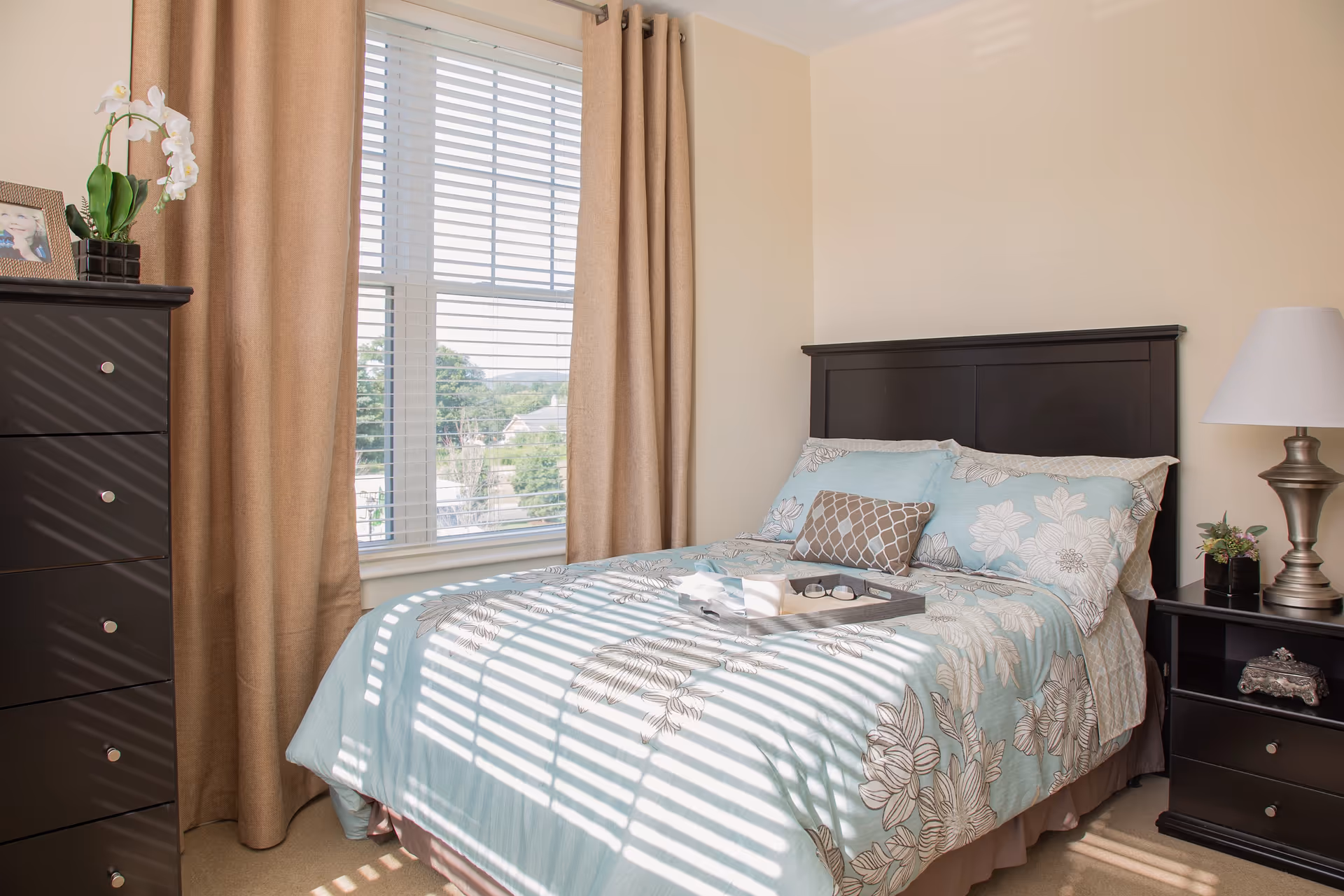 Cozy bedroom featuring a bed with dark headboard, dresser, nightstand, and large window with blinds.