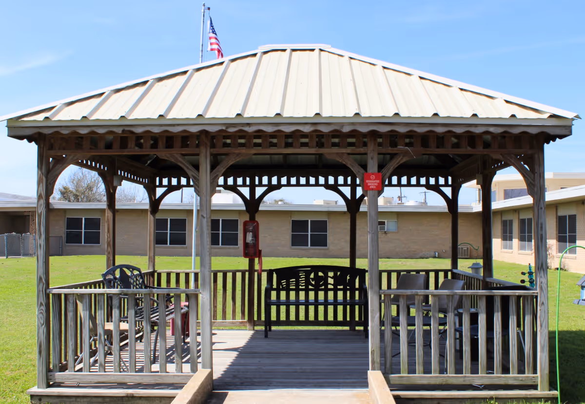 A wooden gazebo with a metal roof situated on a grassy lawn in front of a single-story brick building. Inside the gazebo are several chairs and a bench. An American flag is visible flying on a pole behind the gazebo.