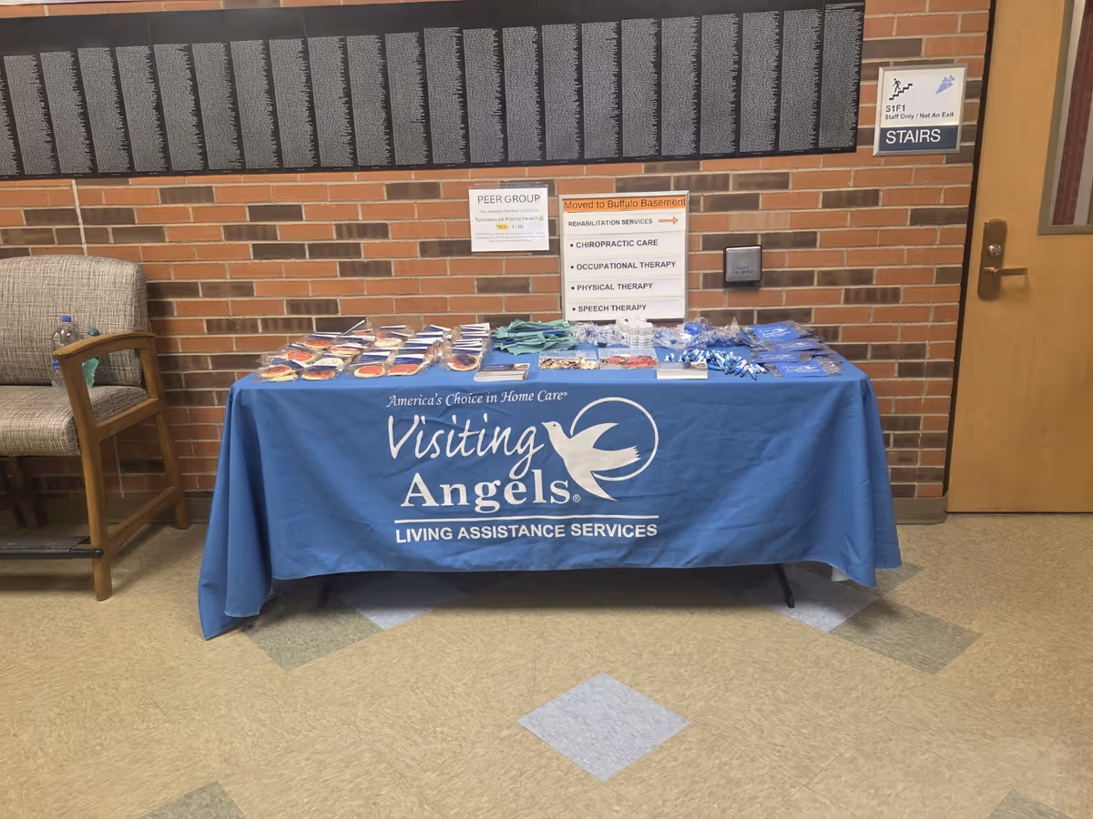 Blue Visiting Angels promotional table with brochures sits in a brick-walled hallway.