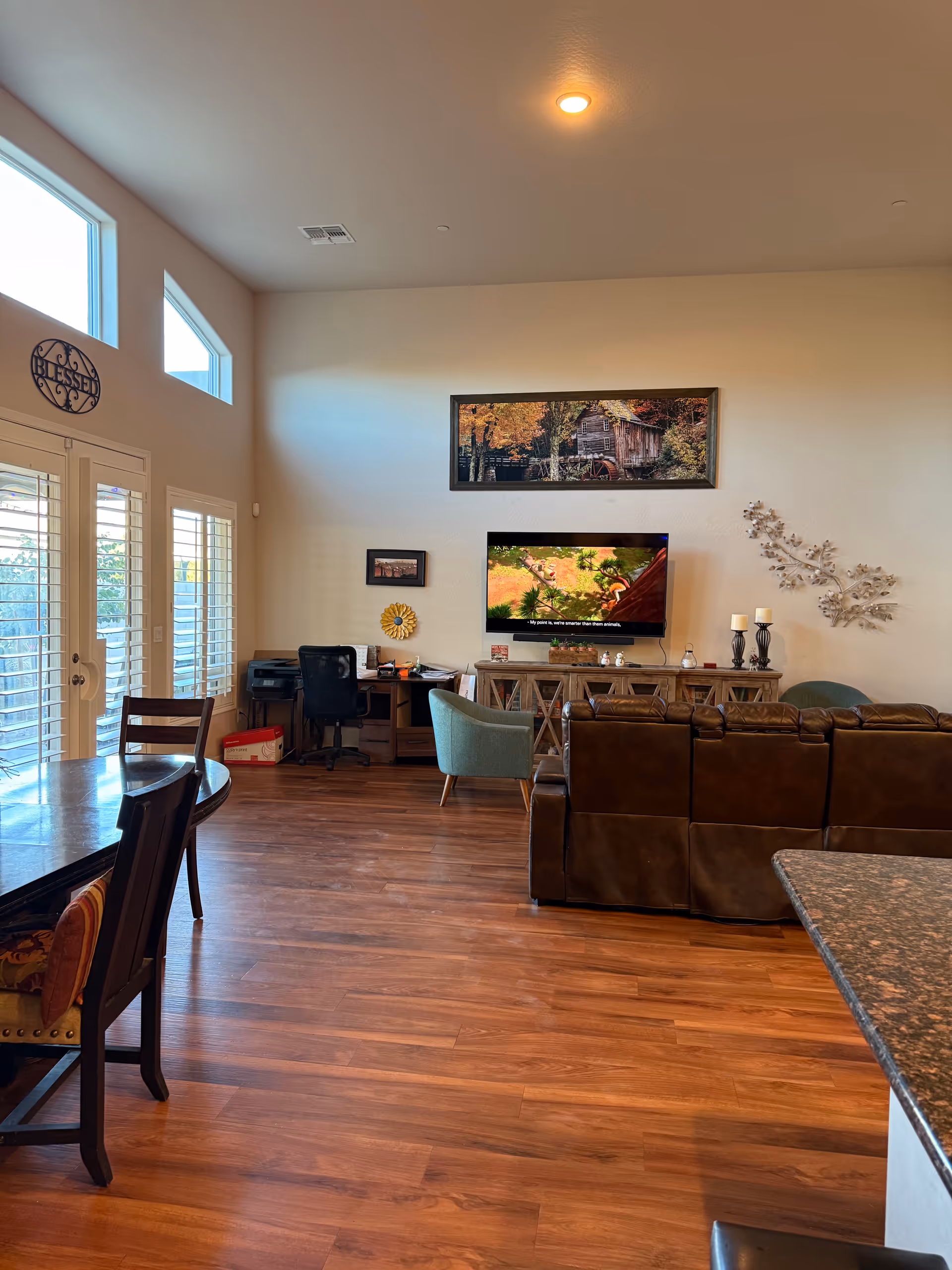 Spacious living room with brown leather seating, TV, desk, artwork, and large windows.