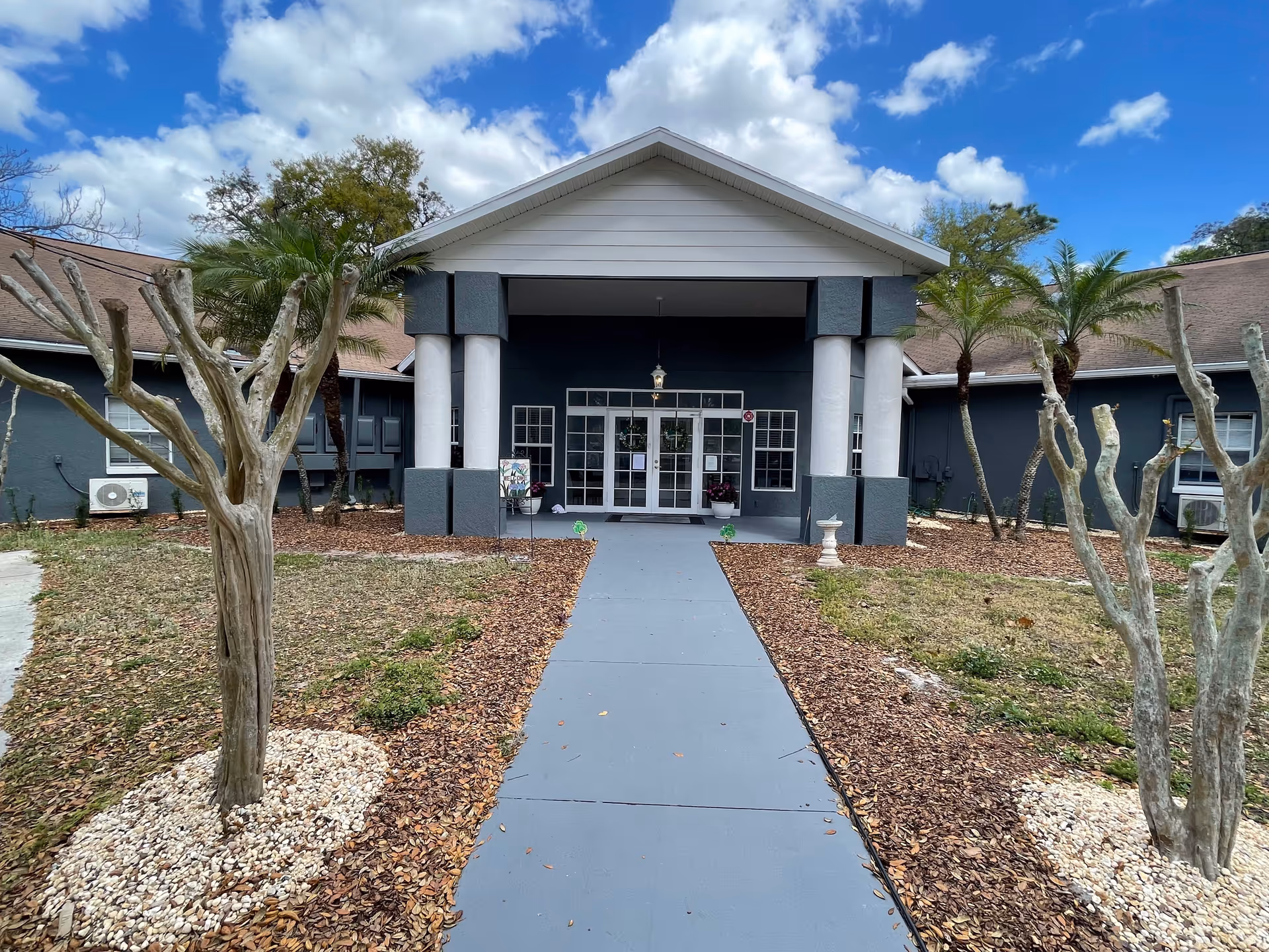 Front entrance of Elysian Gardens Assisted Living Facility with walkway, columns, and palms.
