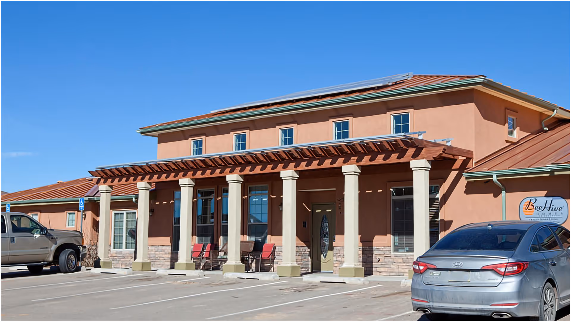 Exterior view of BeeHive Homes Assisted Living facility showing a building with a terracotta-colored facade, stone accents, and a covered entrance supported by columns. There are several chairs on the porch and two cars parked in front under a clear blue sky.