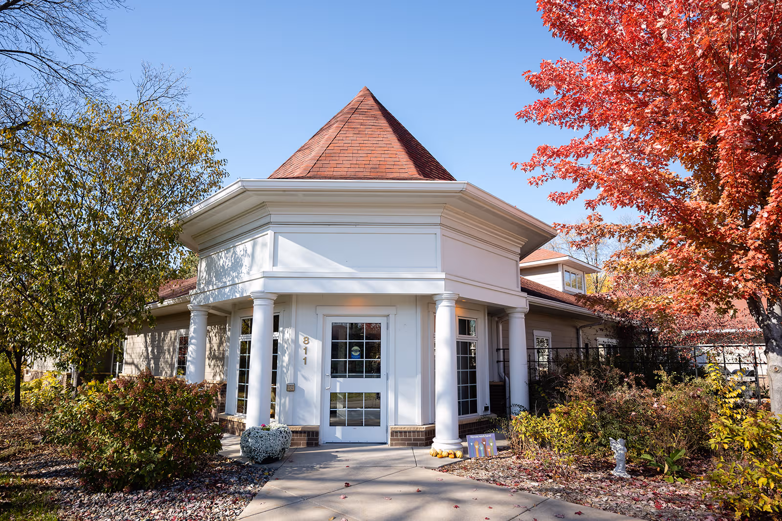 White, columned entry with a red conical roof, framed by autumn trees.