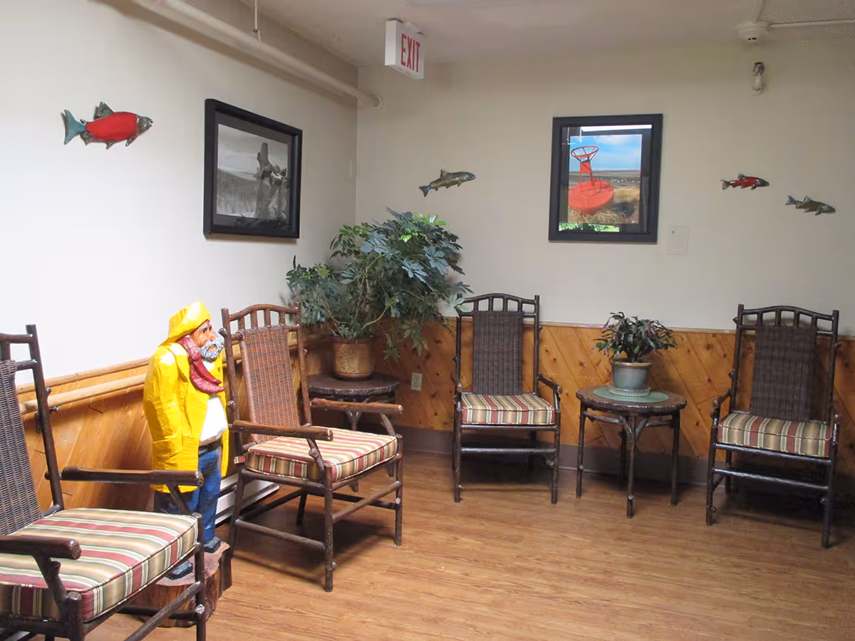 Small interior seating area with wooden chairs with striped cushions, potted plants, wall art and a fisherman statue wearing a yellow raincoat.
