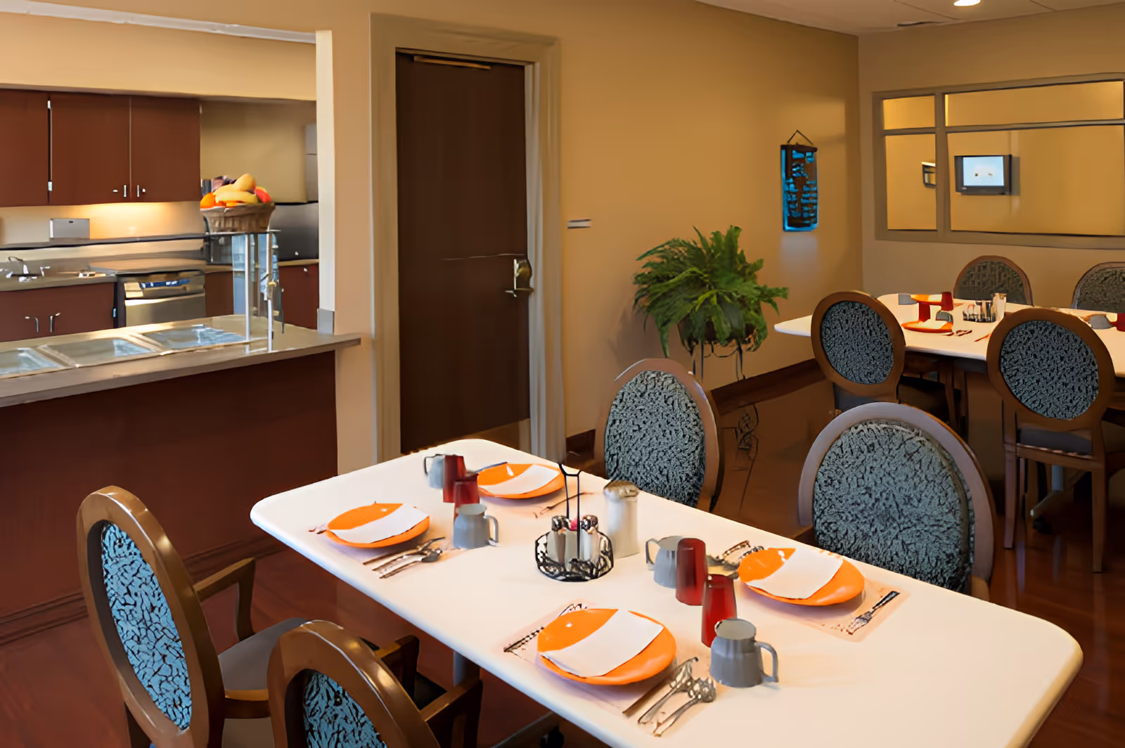 Dining area with tables set with plates and cups, upholstered chairs, and a kitchen counter in the background.