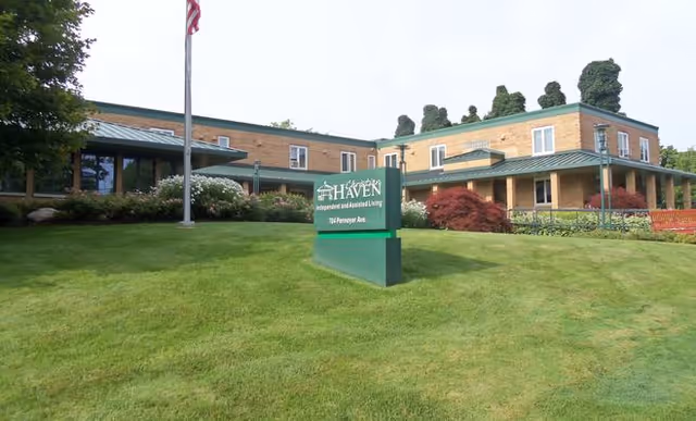 Exterior view of Christian Haven Home, a two-story brick building with a green roof and a covered porch. There is a green sign in front of the building with the facility name and address, surrounded by a well-maintained lawn and landscaping. An American flag is also visible on a flagpole near the sign.