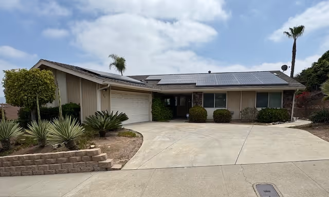 Single-story residential building with a two-car garage, solar panels on the roof, and a driveway. The front yard has various plants and small trees, including palm trees, under a partly cloudy sky.