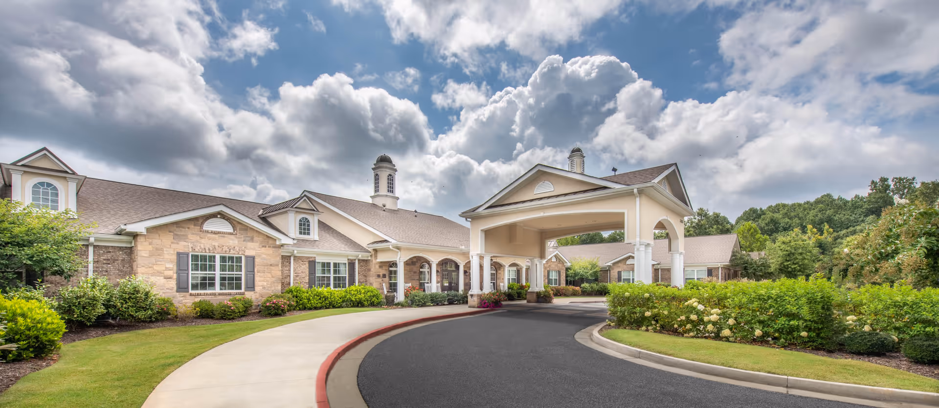 Front entrance of a single-story stone and brick senior living building with a covered porte-cochere and landscaped curved driveway under a cloudy sky.