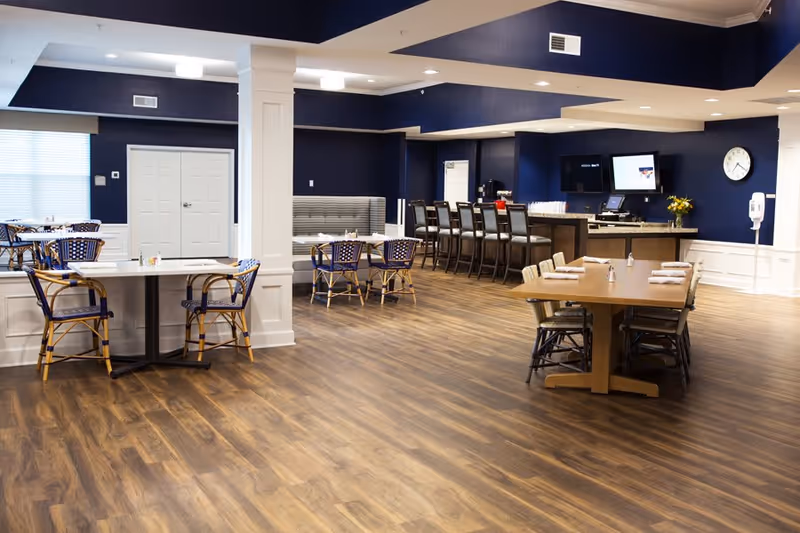 A spacious dining area with wooden flooring and navy blue walls. The room features several tables with chairs, including a rectangular table set with napkins and salt and pepper shakers, and a bar area with high stools. There is a clock on the wall and two monitors behind the bar counter.