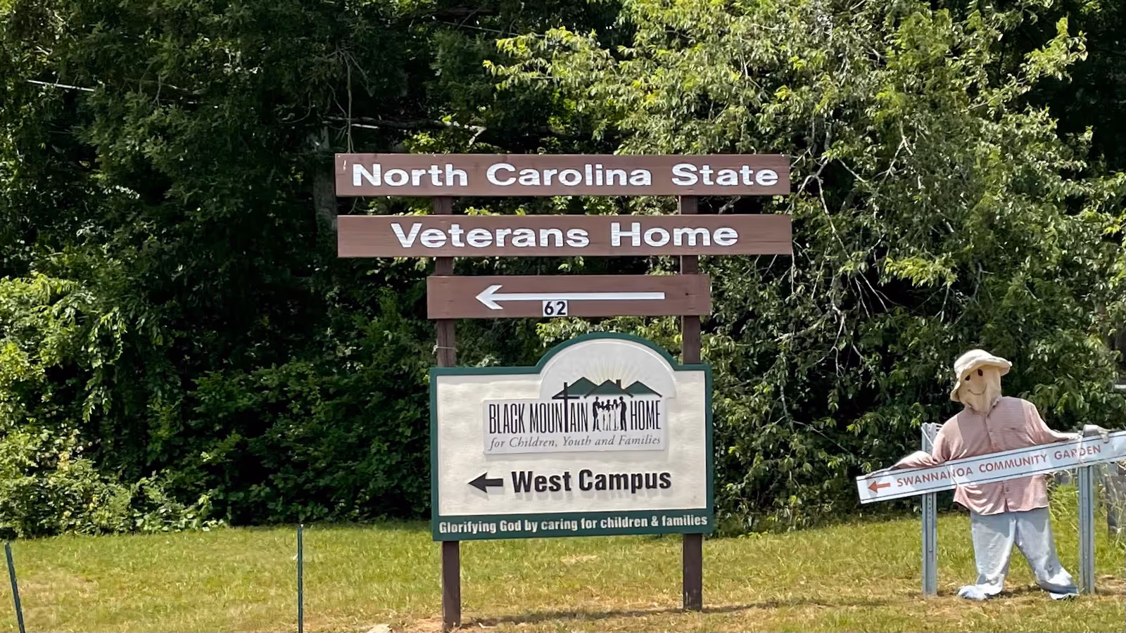 Wooden signs for North Carolina State Veterans Home amid trees, with a scarecrow statue nearby.