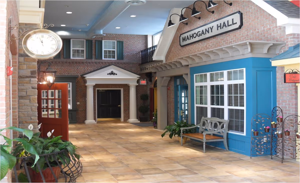 Indoor common corridor styled like a small-town street with storefront facades, a bench, plants and a 'Mahogany Hall' sign.
