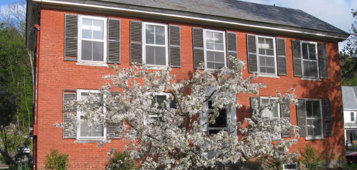 Front exterior view of a two-story red brick building with multiple windows featuring dark shutters. A flowering tree with white blossoms is in front of the building, partially obscuring the entrance door.
