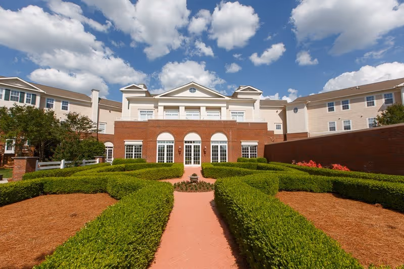 Front view of a brick-faced senior living building with a central walkway through manicured hedges leading to arched glass doors under a blue sky.