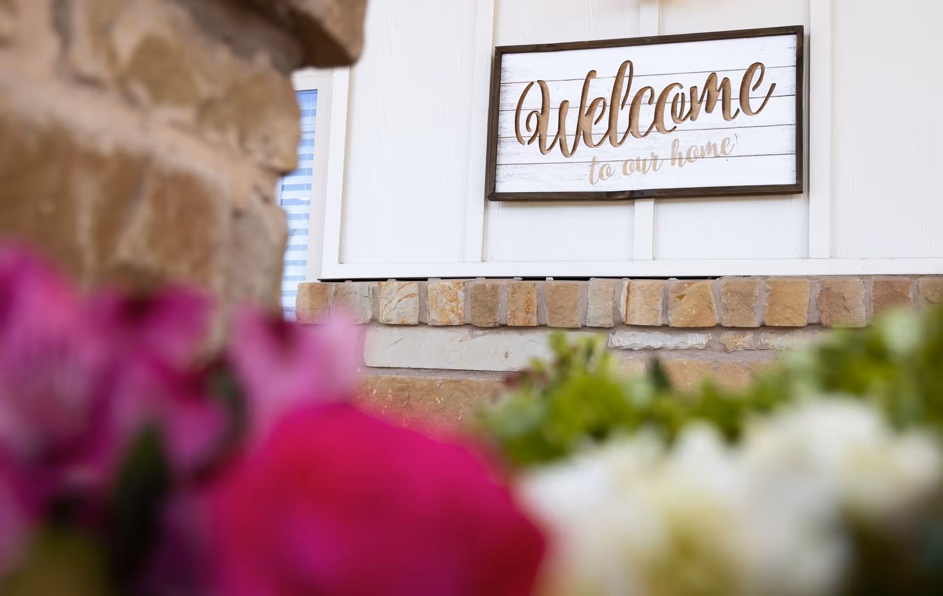 A 'Welcome to our home' sign mounted on a white exterior wall above a brick ledge with blurred flowers in the foreground.