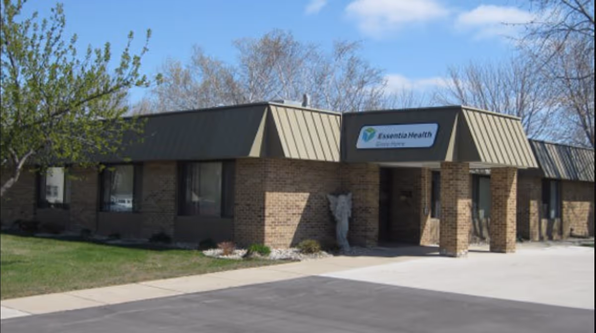 Exterior view of a single-story brick building with a green metal roof and a covered entrance. A sign above the entrance reads 'Essentia Health Grace Home'. There is a small statue near the entrance and some trees and grass surrounding the building under a partly cloudy sky.