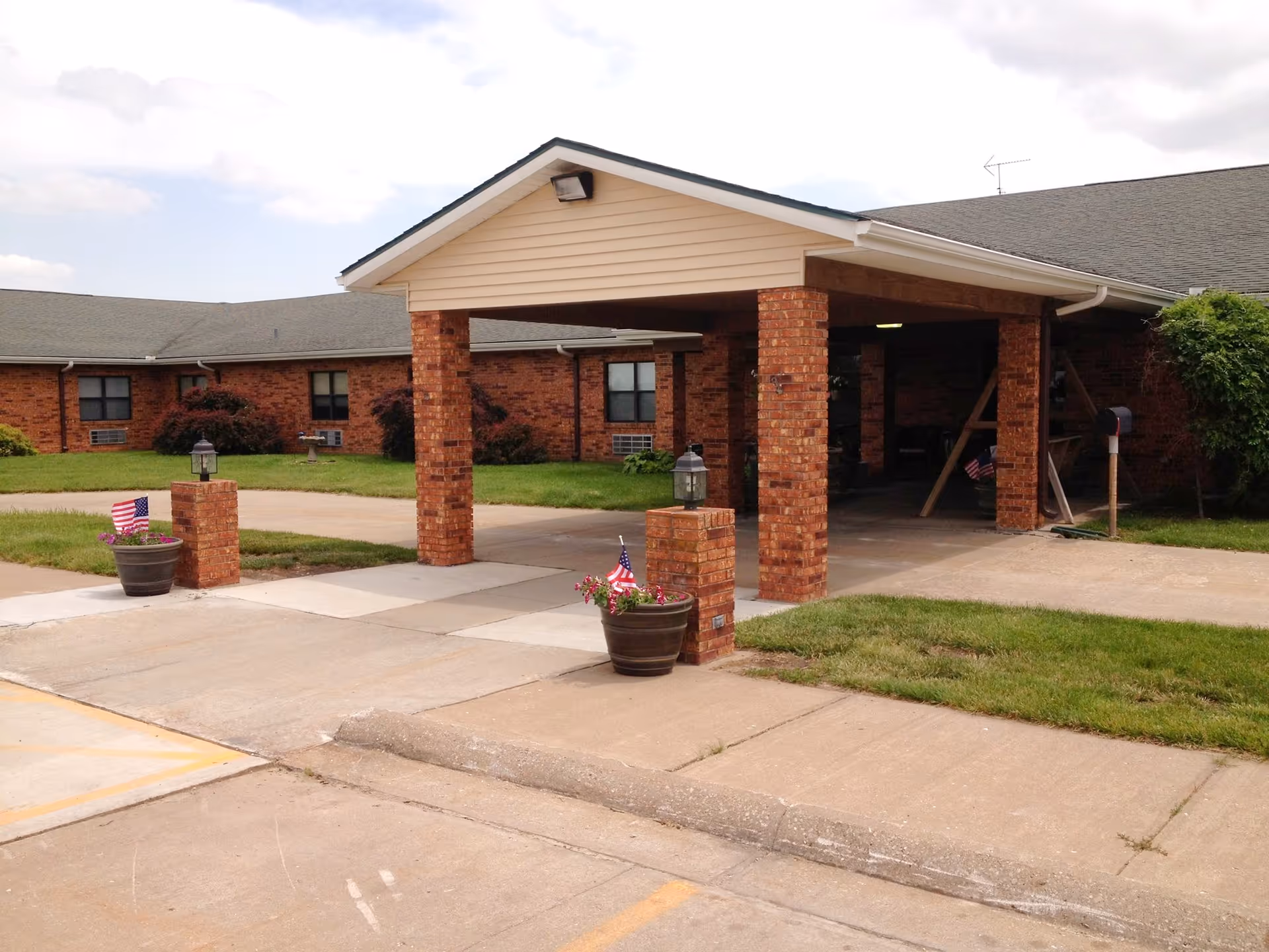 Entrance area of a brick building with a covered drop-off area supported by brick pillars. There are flower pots with small American flags on either side of the entrance, a mailbox to the right, and a grassy lawn surrounding the building.