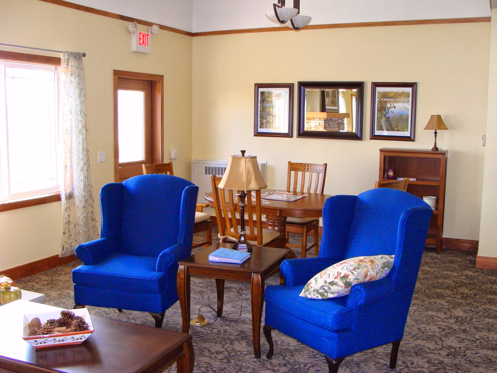 A cozy living room area with two bright blue upholstered armchairs, a wooden side table with a lamp and a book, a wooden dining table with four chairs, framed artwork on the wall, a window with sheer curtains, and a wooden door with an exit sign above it.