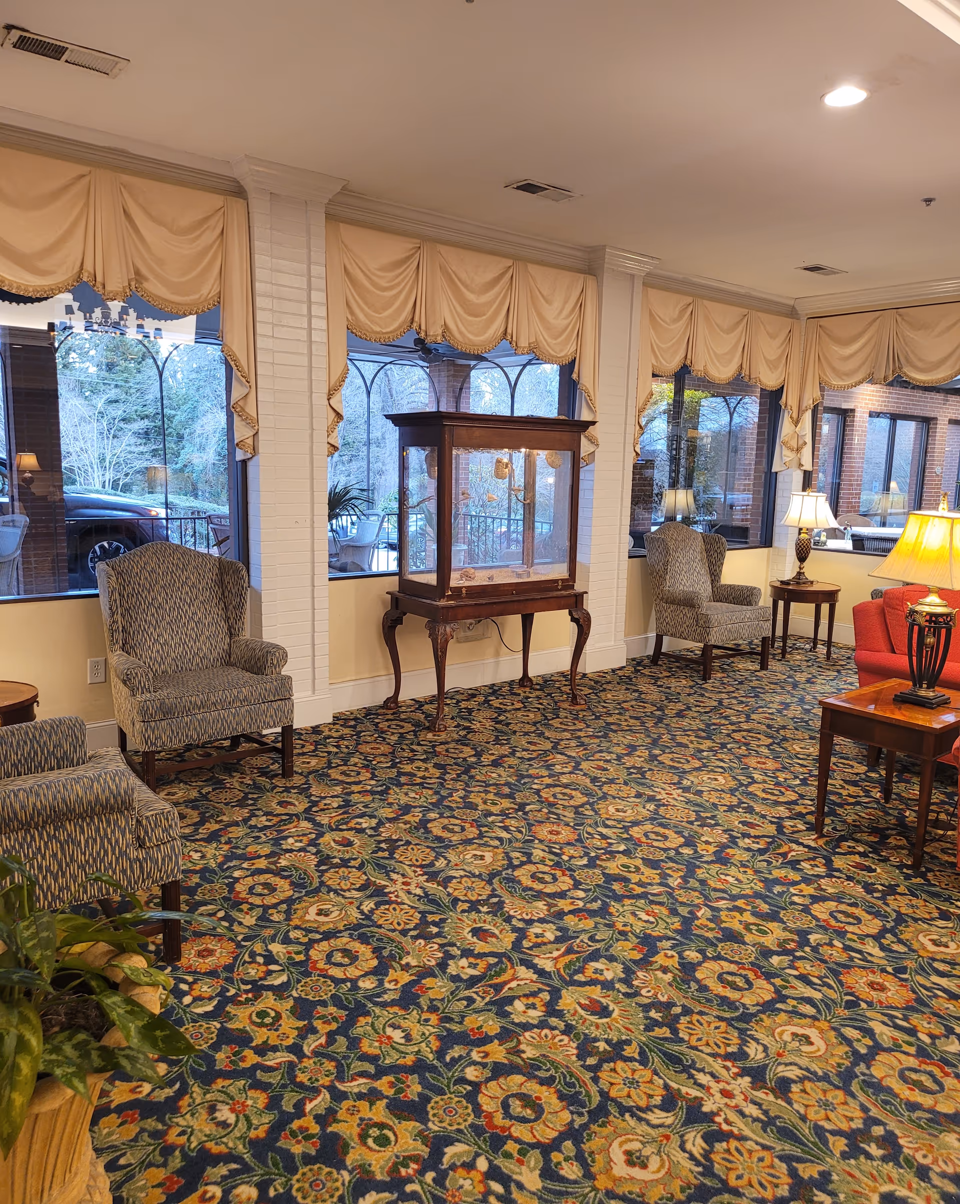 Senior living lobby with patterned carpet, multiple armchairs, lamps, and a glass display cabinet.